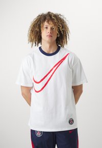 Camiseta blanca de algodón con un gran gráfico del swoosh rojo de Nike, cuello de canalé azul marino y un parche con el logotipo en el lado.