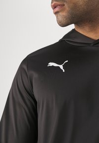 Crna dukserica s kapuljačom izrađena od glatkog, laganog materijala s bijelim skakanjem Puma logotipom na prsima. Jednostavan dizajn, bez uzoraka.