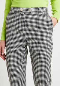 Kalhoty s černobílým houndstooth vzorem a dvěma stříbrnými knoflíky v pase, které jsou nošeny s limetkově zeleným svetrem.