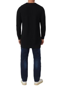 Homme portant un long pull noir, un jean bleu foncé et des baskets blanches, se tenant de dos sur un fond blanc.