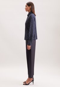Donna di profilo che indossa una blusa blu marina con pois, pantaloni neri su misura e sandali alti neri, in piedi contro uno sfondo neutro.
