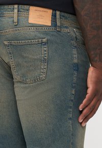 Tamno plave traper hlače s malo izblijedjelim teksturom, koje imaju stražnju džep i smeđu kožnu oznaku s natpisom "JACK & JONES."