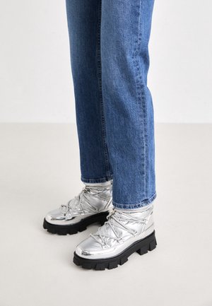 Osoba ubrana w niebieskie dżinsy i srebrne metaliczne sznurowane buty z grubą czarną podeszwą, stojąca na jasnej podłodze.