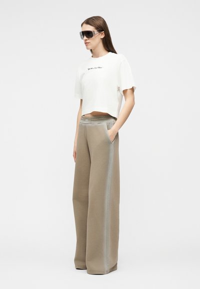 Γυναίκα που φοράει φαρδιά μπεζ παντελόνια με λευκή κατακόρυφη ρίγα, λευκό κοντό T-shirt και μεγάλα μαύρα γυαλιά ηλίου, στέκεται με τα χέρια στις τσέπες.