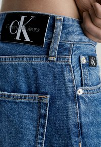 Jeans de denim con un lavado azul oscuro, costuras visibles, una etiqueta negra con "Calvin Klein Jeans" y un pequeño logotipo en el bolsillo trasero.