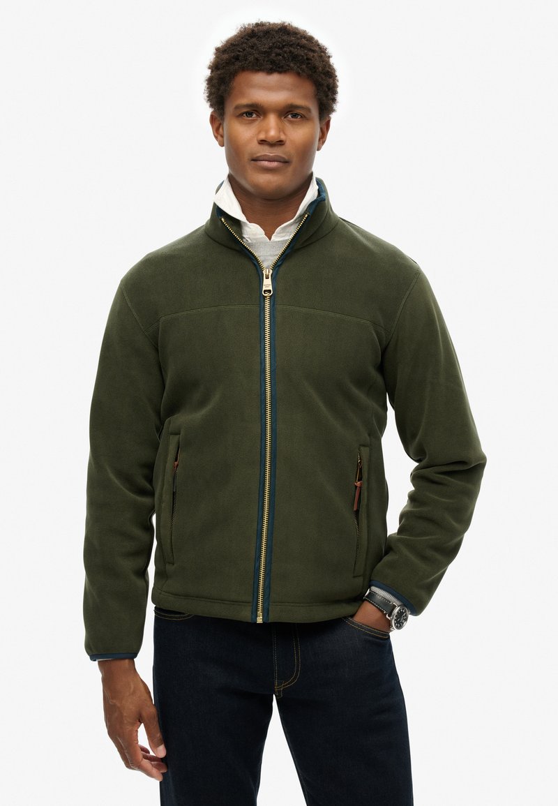 Giacca in pile verde con cerniera blu, dotata di tasche laterali, colletto alto e tessuto testurizzato; abbinata a jeans scuri.