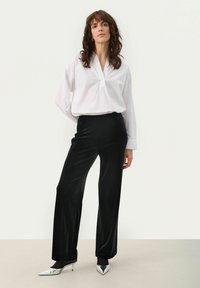 Bluză albă cu guler și o placă cu jumătate de butoane, combinată cu pantaloni largi din catifea neagră și tocuri argintii ascuțite. Texturi netede.