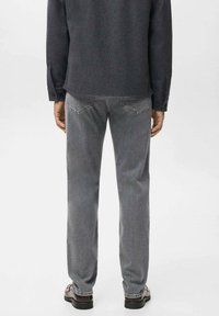 Man in grijze spijkerbroek, donker shirt met lange mouwen en bruine schoenen, staand met zijn rug naar de camera tegen een effen achtergrond.