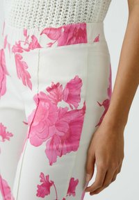 Pantalon blanc avec grand motif floral rose, présentant une texture lisse et une coupe ajustée, montré à la taille avec une main posée dessus.