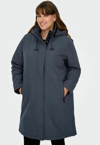 Smaidoša sieviete, tērpusies garā tumši pelēkā ziemas mētelī ar kapuci, rokām kabatās ar rāvējslēdzējiem uz vienkārša fona.