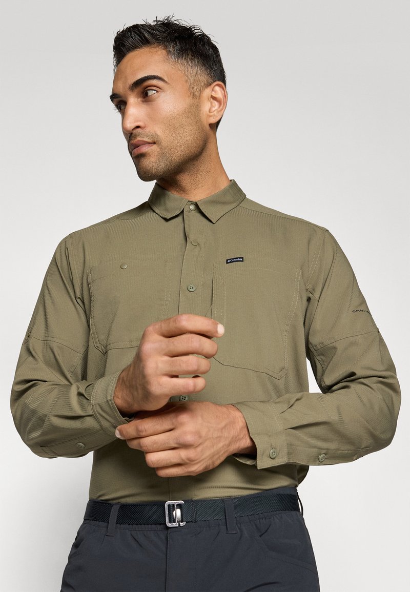 Langärmliges, olivgrünes Hemd mit Button-Down-Kragen, zwei Fronttaschen und strukturiertem Stoff; kombiniert mit dunklen Hosen und einem Gürtel.