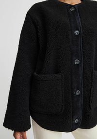 Giacca in fleece nera con superficie testurizzata, patta frontale in corduroy e due tasche anteriori. Presenta grandi bottoni e un collo rotondo.