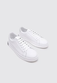 Zapatillas blancas de cuero con una textura suave, punta redonda y diseño con cordones. Presentan un detalle gris en el talón. Estilo minimalista y moderno.