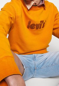 Personne portant un sweat-shirt Levi's orange vif superposé à une chemise à col et un short en jean bleu clair, assise les jambes écartées.