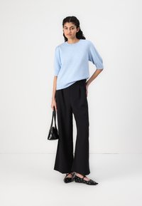 Top de malha azul claro com mangas curtas, combinado com calças pretas de perna larga. Os acessórios incluem uma pequena mala preta e sapatilhas pretas com tachas.