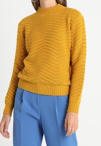Gestickter Senfgelber Pullover mit hohem Kragen, der ein Wellenmuster und gerippte Bündchen und Saum aufweist, kombiniert mit blauen Hosen.