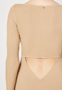 Haut long à manches ribbé couleur tan avec un col rond et une découpe au dos, fabriqué à partir d'un tissu extensible. Étiquettes visibles au niveau du col.