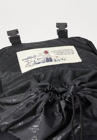 Svart nylon ryggsekkinnside med snøring, trykte illustrasjoner og en beige etikett med tekst og grafisk design.
