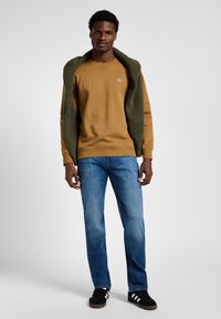 Mustard yellow sweatshirt with a green knit collar, paired with blue denim jeans and black sneakers with white stripes. Model stands against a light background.