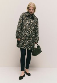 Žena vo februárovom kabáte s leopardím vzorom, čiernych nohaviciach a balerínach, drží zelenú kabelku a stojí pred jednoliatym svetlým pozadím.