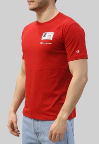 T-shirt in cotone rosso con scollatura a girocollo, dotata di una toppa bianca con logo e testo sul petto e marchio Champion sulla manica.