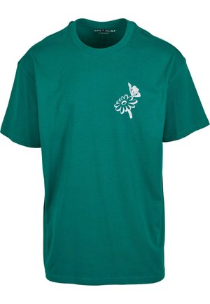 Teal puuvillapaita, jossa on valkoinen kukka- ja perhoskuvio vasemmalla puolella. Lyhyet hihat ja pyöreä kaula-aukko.