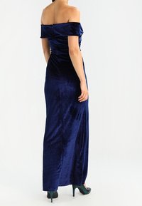 Mörkblå sammet klänning med avskuren axel och figursydd silhuett, golvlång och med subtil lyster; matchad med mörkt teal klackar.