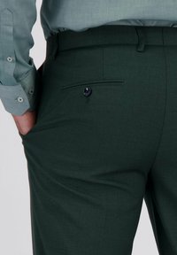 Donkergroene broek met een gladde textuur, voorzien van een achterzak met een blauwe knop en een op maat gemaakt pasvorm. Vergezeld van een lichtblauw overhemd.