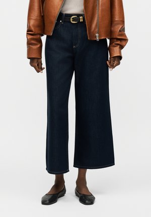 Άτομο που φοράει φαρδιά μπλε σκούρα τζιν, καφέ δερμάτινο μπουφάν, μαύρη ζώνη και μαύρες μπαλαρίνες σε απλό φόντο.