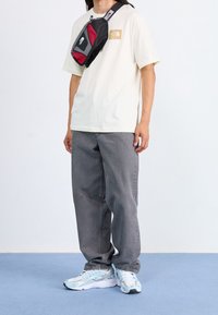 Svart och röd axelväska, beige t-shirt med logotyp, grå vida byxor och ljusblå sneakers med grå detaljer på en blå bakgrund.