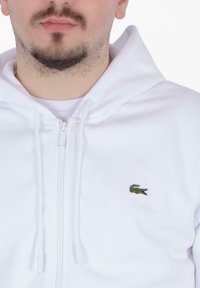 Felpa bianca con zip realizzata in tessuto morbido, con un piccolo logo di coccodrillo verde sul lato sinistro del petto. Include coulisse sul collo.