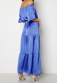 Robe longue en tissu bleu lisse, style épaules dénudées, avec des manches bouffantes et un ourlet à volants. Associée à des sandales à talons bleu clair.