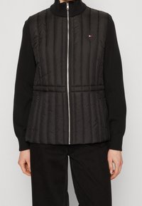 Gilet imbottito nero con texture a coste verticali, colletto alto e zip argentata. Presenta il logo del marchio e tasche laterali con zip. Indossato sopra una maglietta nera.