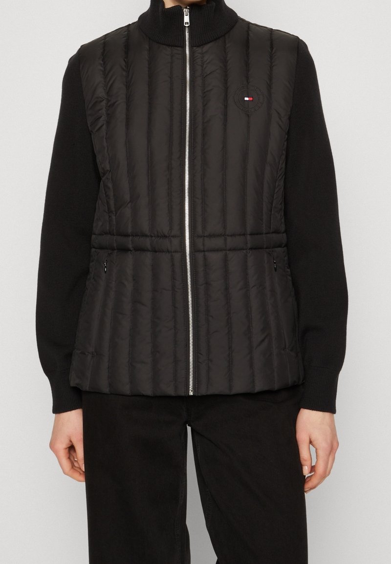 Gilet imbottito nero con texture a coste verticali, colletto alto e zip argentata. Presenta il logo del marchio e tasche laterali con zip. Indossato sopra una maglietta nera.