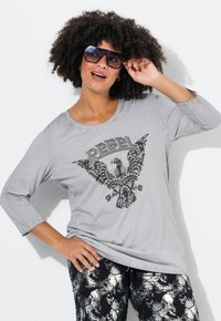 Graues Longsleeve-Shirt mit schwarzem Adler-Design und dem Text "REBEL". Das Modell trägt dunkle gemusterte Hosen und übergroße Sonnenbrillen.