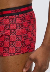 Κόκκινα boxer briefs με σχέδιο πλέγματος και "HUGO" σε μαύρο, μαύρη ζώνη μέσης με κόκκινα γράμματα, κατασκευασμένα από ελαστικό ύφασμα, εφαρμοστή γραμμή.