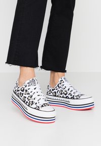 Piedi che indossano sneakers Converse con stampa leopardata bianca e suole a piattaforma con righe rosse, bianche e blu, abbinate a pantaloni neri a metà polpaccio.