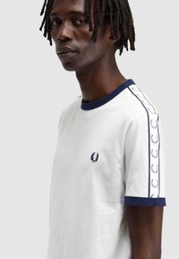 Camiseta de algodón blanca con cuello y puños de canalé en azul marino. Detalles notables incluyen un logo en el pecho y rayas decorativas en las mangas.