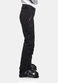 Schwarze Skihose aus flexiblem Material mit schlankem Schnitt, ausgestattet mit einer Reißverschlusstasche und einem gestickten Logo nahe der Hüfte. Getragen mit schwarzen Skischuhen.