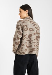 Gebreide trui in beige met bruine abstracte patronen. Ronde hals, ontspannen pasvorm en gemêleerd materiaal. Gecombineerd met zwarte, high-waisted broek.