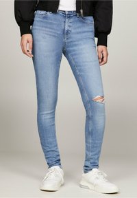 Persona vestida con unos pantalones ajustados rasgados de color azul claro, zapatillas blancas, una chaqueta negra y una blusa blanca, de pie sobre un fondo neutro.