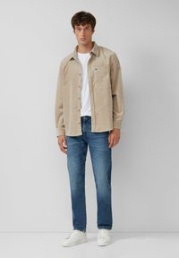 Chemise en velours côtelé beige avec une poche avant, superposée sur un t-shirt blanc; associée à un jean bleu et des baskets blanches. Look simple et décontracté.