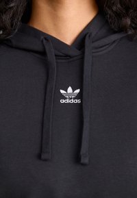 Felpa nera con cappuccio, tasca frontale, coulisse e logo trefoil Adidas bianco sul petto. Realizzata in morbido tessuto di cotone.