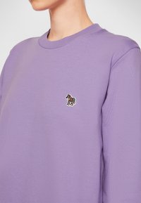 Osoba v levandulovém svetru s kulatým výstřihem, na levé straně hrudi s malým vyšitým barevným zebra.