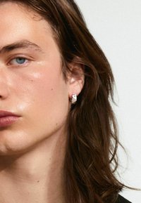 Orecchino a cerchio d'argento con una pietra trasparente, con una finitura lucida. Il modello ha lunghi capelli castani e un tono di pelle neutro.