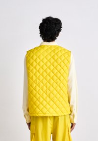Gilet matelassé jaune au design sans manches, avec un motif en losanges. Porté par-dessus une chemise de couleur claire et associé à un pantalon jaune.
