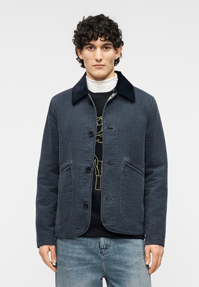 Veste matelassée bleu marine avec col en velours côtelé, six boutons sur le devant et deux grandes poches latérales, portée par-dessus un t-shirt noir et un jean bleu clair.