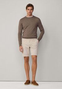 Brązowy sweter dzianinowy, beżowe szorty i brązowe mokasyny. Model stoi z jedną ręką w kieszeni, biała koszulka pod okresem widoczna przy dekolcie.