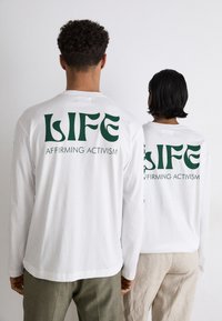 Weiße Langarmshirts mit großen grünen Aufschriften, die "LIFE" und "AFFIRMING ACTIVISM" auf der Rückseite zeigen. Aus glattem Baumwollstoff.