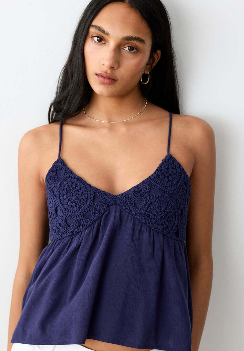 Femme aux longs cheveux noirs portant des boucles d’oreilles créoles et un haut sans manches bleu marine avec des détails au crochet sur la poitrine.